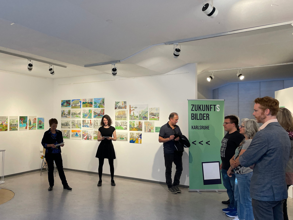 Leute betrachten die Ausstellung "Zukunftsbilder Karlsruhe" in einem Ausstellungsraum, der an eine Galerie erinnert.